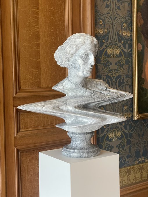 A marble bust of a woman with an elegant updo displayed on a white pedestal, featuring a dramatic 'glitch' or 'melting' distortion effect around its base, set against an ornate wood-paneled wall with patterned wallpaper.