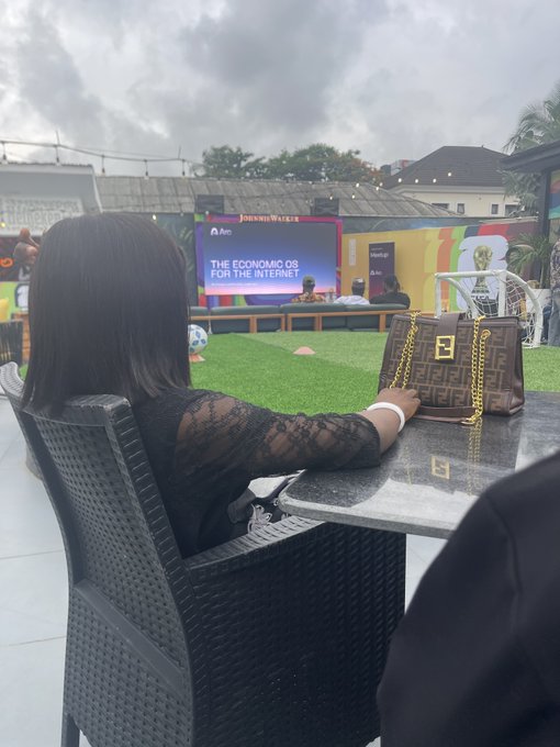 A woman with shoulder-length black hair, wearing a black lace top, sits at a marble table with a designer monogram handbag, watching a stage presentation displaying "Joanna Walters" and "The Economic OS for the Internet," capturing the Arc community event experience in Lagos.