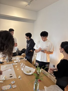 A group of young people gather around a wooden table in a white room, with one person in a white t-shirt looking down at their phone while others chat and interact with items on the table.