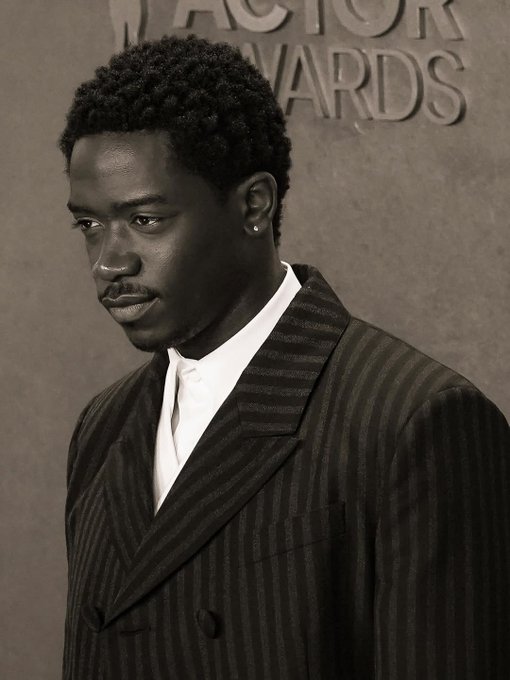 A portrait of a man in a dark pinstripe suit at what appears to be the Actor Awards, based on the text in the background.
