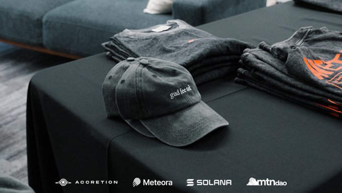 A black cap embroidered with "gnufacet" rests on a black table alongside stacked gray Meteora t-shirts featuring orange branding, with logos for Accretion, Meteora, Solana, and mtndao displayed at the bottom—showcasing the exclusive on-site merchandise available at the mtndao Demo Day event.
