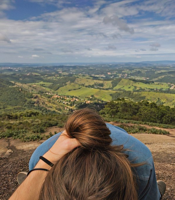 A view from behind of a person with brown hair sitting on a ledge, looking out over a vast landscape of green rolling hills under a cloudy blue sky.