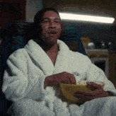 The image shows an actor wearing a white robe, sitting down with food in his hands while looking at something off-camera. The background appears to be a room or a set that resembles a living space. There are two clocks visible in the scene - one on the left side and another on the right side of the frame. A bowl is also present near the actor's hand, possibly containing food.
