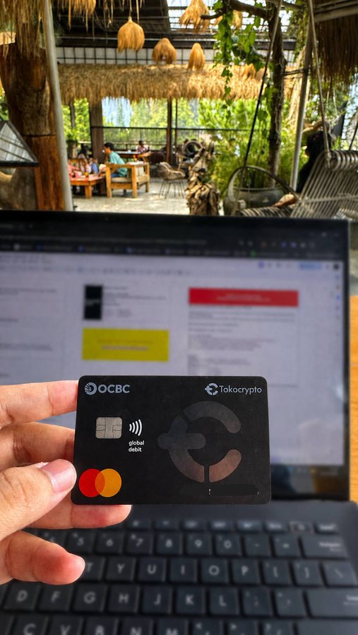 A hand holds a black OCBC debit card with a Tokocrypto logo and a red Mastercard logo in front of an open laptop, with a thatched-roof cafe visible in the background.