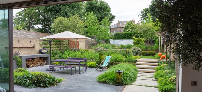 A landscaped backyard garden viewed from inside, featuring a stone patio with a dining set under a white umbrella, a built-in fireplace, manicured plants, and a stone path leading up steps.