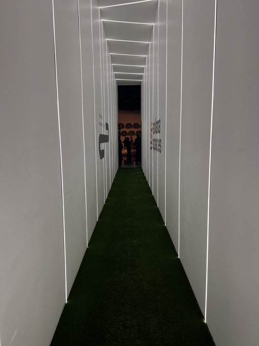 A long, narrow corridor lined with white panels and LED strip lighting, featuring artificial green turf flooring and people visible at the far end.