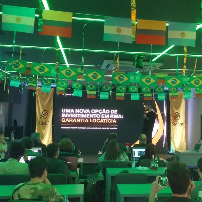 A conference or event space decorated with numerous Brazilian and Argentinian flags hanging from the ceiling, where attendees sit at desks facing a screen displaying a Stellar presentation titled 'UMA NOVA OPÇÃO DE INVESTIMENTO EM RWA: GARANTIA LOCATÍCIA.'