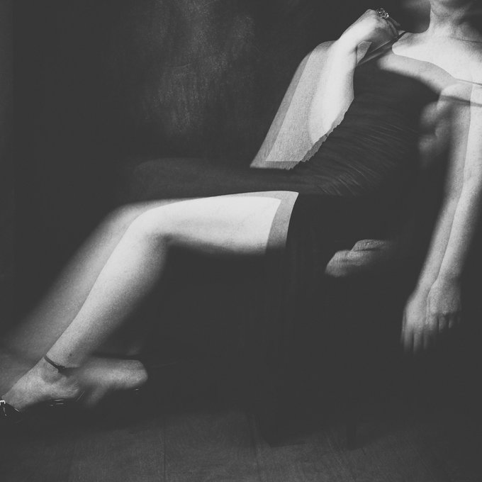 Black and white photo of a woman in a short black dress seated, her legs crossed and extended, with dramatic lighting.