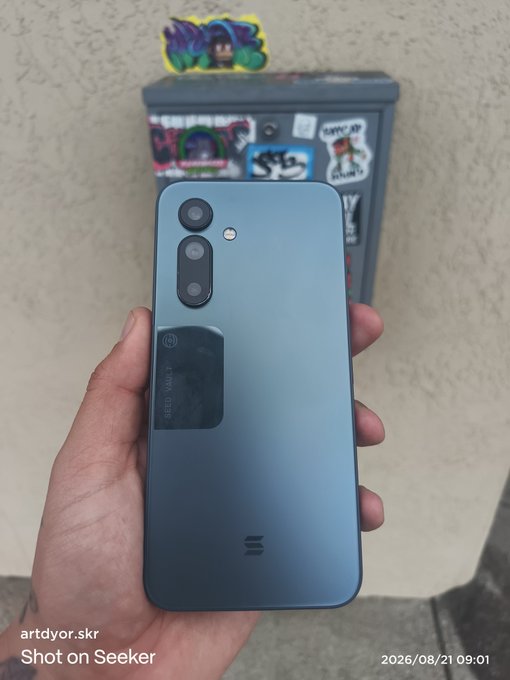 A person's hand holds up a blue Xiaomi smartphone showing its back cameras; an electrical box covered in stickers is visible in the background, with a watermark reading 'artdyor.skr Shot on Seeker 2026/08/21 09:01'.