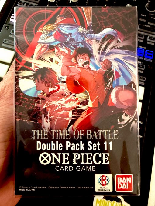 A hand holds a sealed "One Piece Card Game" booster box titled "THE TIME OF BATTLE Double Pack Set 11," featuring anime artwork of Luffy and other characters, with Bandai and Bandai Card Games logos, made in Japan, photographed over a MIDI keyboard.