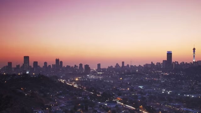A panoramic cityscape at sunset or sunrise shows a skyline with skyscrapers, a tall communications tower on the right, and pink-to-orange gradient skies over an urban area.
