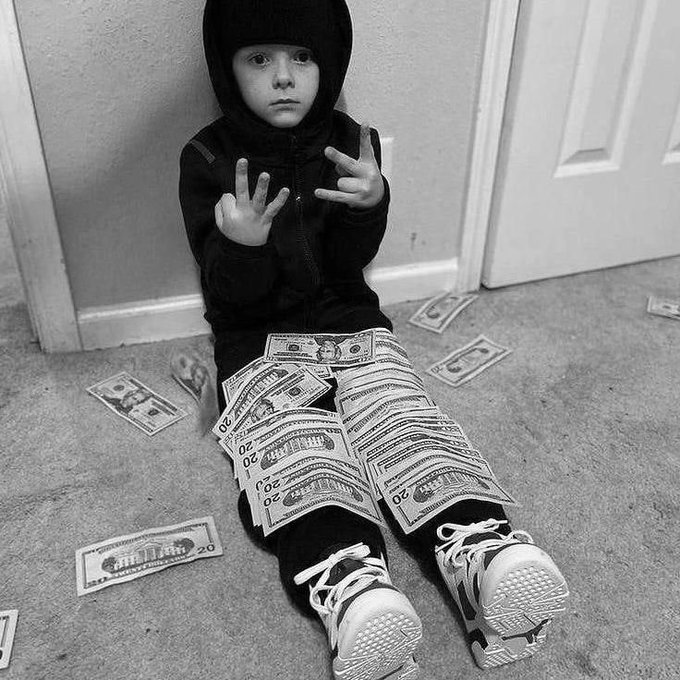 A black-and-white photograph features a young child in a dark hoodie and sneakers sitting on the floor, draped with twenty-dollar bills and making hand gestures.