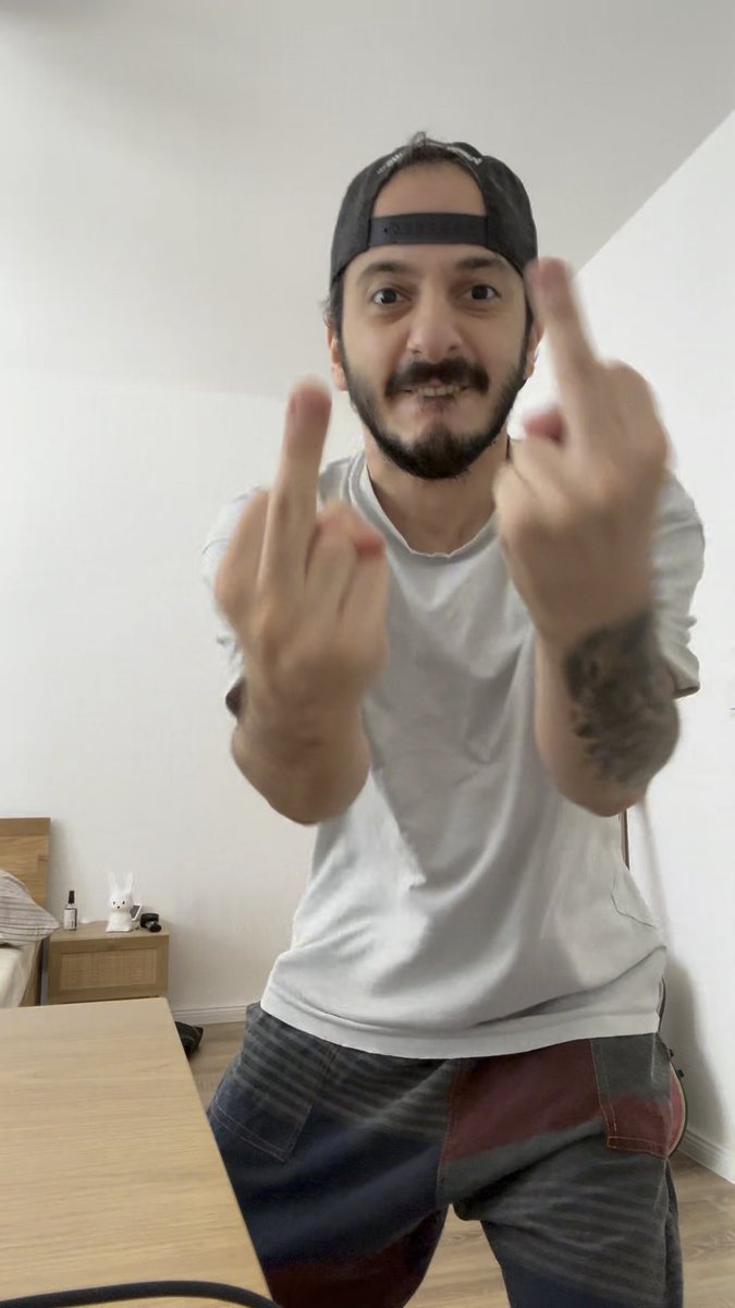 A bearded man wearing a backwards black cap and white t-shirt smiles and gestures toward the camera with both middle fingers raised in a room with white walls.