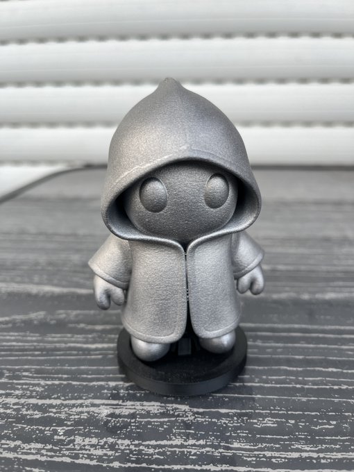 A small, gray, hooded figurine resembling a cute chibi character with large round eyes, displayed on a dark wooden surface.