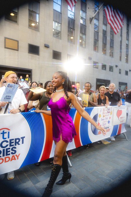 A woman in a vibrant pink dress and black boots smiles while walking past a crowd behind barriers with signs reading 'AYRA STARR' and 'CONCERT STORIES' during a public event.