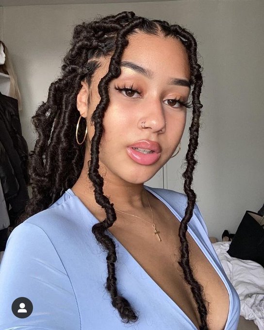 Selfie of a woman with long, twisted locs hairstyle, wearing a light blue top, gold hoop earrings, and a small cross necklace.