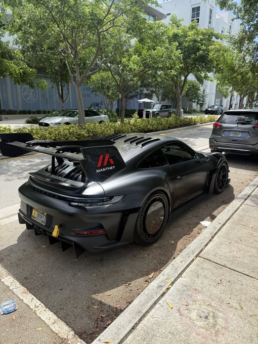 A matte black Porsche 911 GT3 RS with a large rear wing is parked on a Miami street, featuring "MANTHEY" branding on the wing and a yellow license plate reading "D 2277." The post "5 bitcoin or this car?" highlights the car's hefty price tag, which rivals the value of 5 BTC, pitting a tangible luxury asset against digital currency.