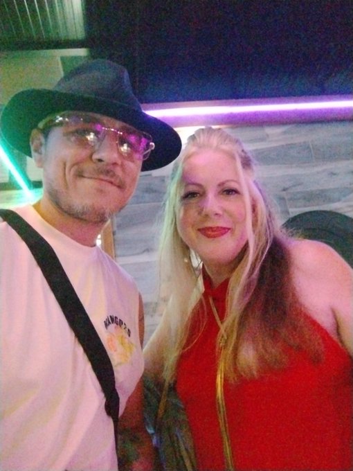 A close-up photo of a smiling man wearing a black hat and glasses, standing next to a blonde woman in a red top in a setting with colorful neon lights.