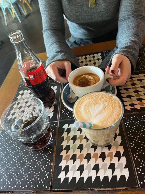 A person in a gray sweater sits at a café table with a Coca-Cola glass bottle and two coffee drinks—a latte with leaf art and a smaller espresso-based drink—on a houndstooth-patterned table, enjoying a morning coffee break to start the day.
