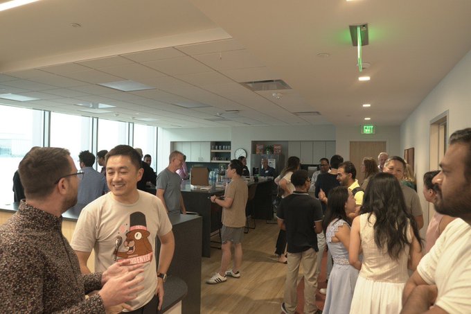 A crowded indoor networking event with people mingling in a modern office space with large windows, where a smiling man in the foreground wears a t-shirt featuring a cartoon hot sauce bottle character.