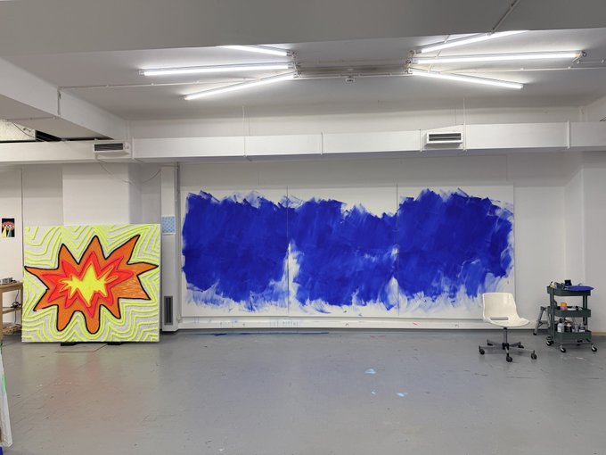 An artist's studio space with bright fluorescent ceiling lights, featuring a large canvas leaning against the wall painted with a vibrant yellow and red starburst/explosion design next to a wall covered in abstract blue paint strokes, with a white rolling chair and art supply cart nearby.