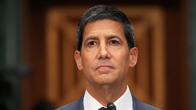 A close-up portrait of a man with short dark graying hair wearing a dark suit, appearing to be in a formal governmental or institutional setting.