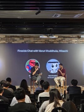 Two people sit on stools on a stage during a 'Fireside Chat with Varun Vruddhula, Hibachi' in front of a seated audience, with their photos displayed on the screen behind them.