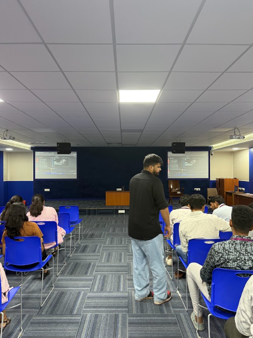 An indoor classroom scene with students seated in blue chairs facing a projection screen, while a man in a black shirt stands in the aisle.
