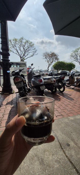 A hand holds a glass of dark soda beneath umbrellas, with parked motorbikes and a waterfront visible in the background.