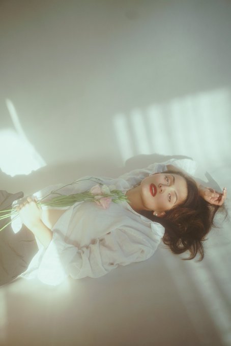 A young woman lies on a white bed, holding pink flowers, wearing a white shirt with soft natural light streaming across the scene.