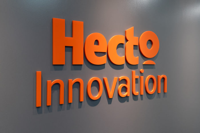 An office signage photo showing the company name 'Hecto Innovation' in large orange 3D letters mounted on a gray wall.