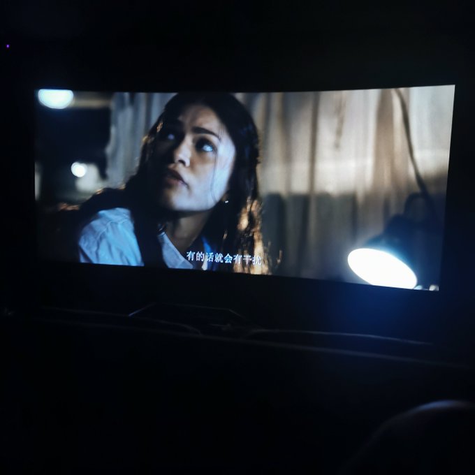 A person watching a movie on a screen showing a woman in a dimly lit room with Chinese subtitles at the bottom.