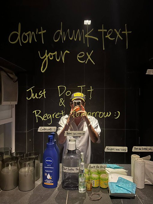 A bathroom mirror with handwritten yellow text reading 'don't drunk text your ex Just Do It & Regret Tomorrow :)' along with humorous labels on various toiletries, as a person takes a selfie reflection.