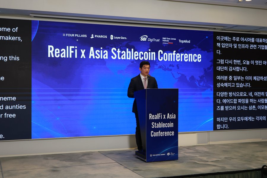 A man in a suit stands at a podium at the 'RealFi x Asia Stablecoin Conference', with sponsors including Four Pillars, Pharos, LayerZero, SBI DigiTrust, S&P Dow Jones Indices, and TopMed displayed on the screen behind him.