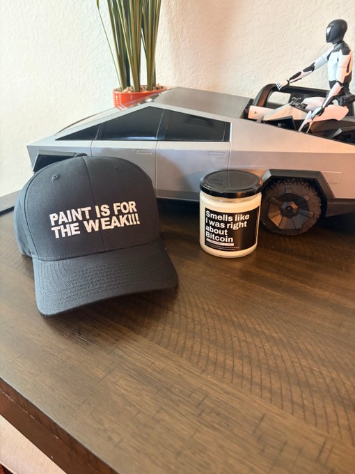 A still life on a wooden table showing a black cap reading 'PAINT IS FOR THE WEAK!!', a candle with text 'Smells like I was right about Bitcoin', and a Cybertruck toy with a small figurine, with a plant in the background.