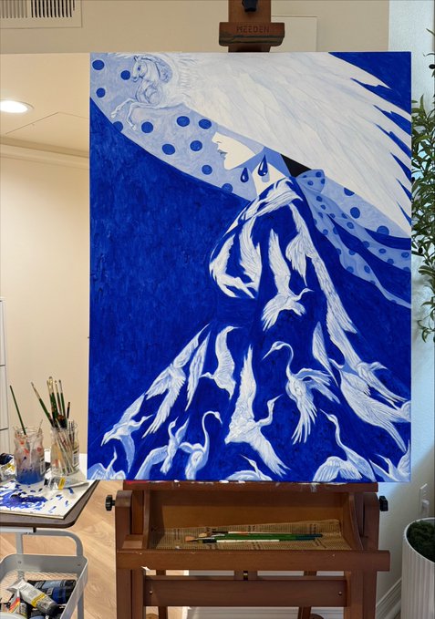 The same painting as image 6 — a blue-toned artwork on a wooden easel showing a figure with flowing white hair, a polka-dot headpiece, and descending white birds, surrounded by art supplies.