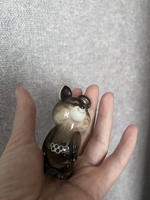 A small, glossy brown ceramic bear figurine with a white snout and the number '1980' on its chest rests in the palm of a person's hand.