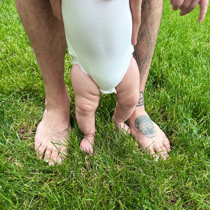 The image shows a man holding his baby's feet while standing in a field of green grass. The baby is wearing a white onesie with a skull tattoo on their foot. In the background, there are two more people partially visible, but they don't seem to be involved in this scene. The post appears to be about teaching children about solana and decentralized finance through a fun and engaging activity involving toys like pokémon and stablecoins.