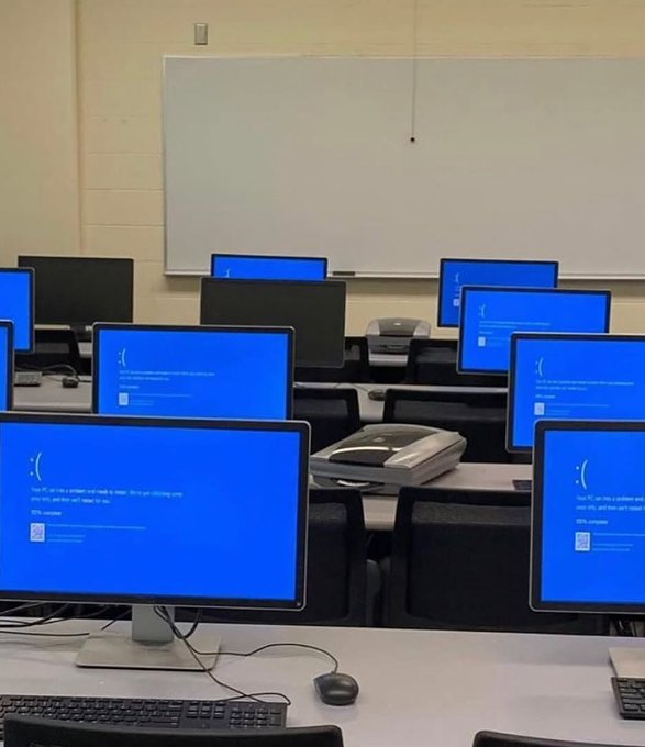 A computer lab or classroom filled with rows of Dell monitors all displaying the same blue Windows error screen with a sad face emoticon and troubleshooting text, with a whiteboard visible on the wall behind them.