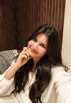 A smiling woman with long dark hair wearing a white top poses indoors, resting her chin on her hand in a cozy setting with wooden walls.