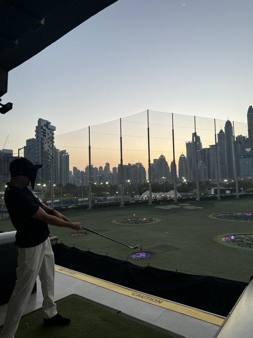 A man wearing a cap and mask stands in a golf driving range bay at dusk, holding a club with a city skyline visible behind the protective netting.