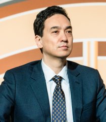 A man in a dark blue suit, white shirt, and patterned blue tie stands against a blurred orange background.