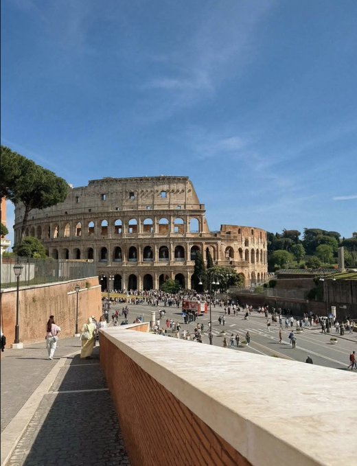 The Colosseum in Rome under a clear blue sky, with tourists walking along the street in the foreground.