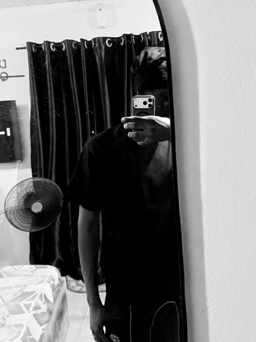 A black and white photo of a person taking a mirror selfie while holding a phone, wearing a black shirt, with a fan and curtains visible in the reflection.