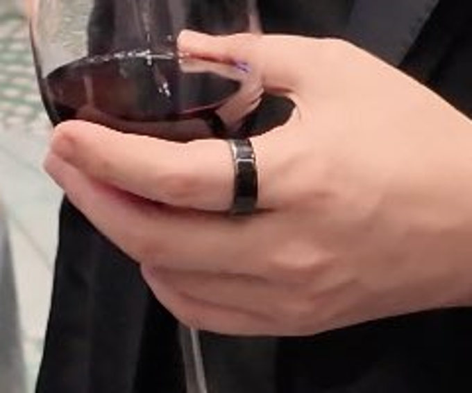 A close-up shows a hand holding a glass of red wine, featuring a wide black band ring on the middle finger and purple nail polish.