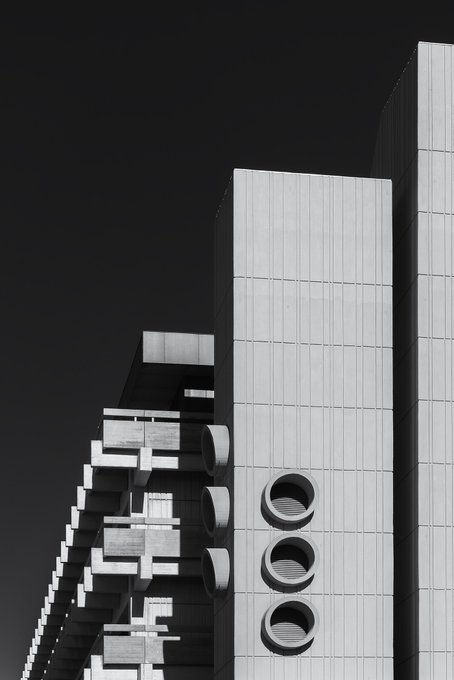 A striking black and white photograph of a brutalist concrete building with circular windows and protruding geometric architectural elements against a stark black sky.
