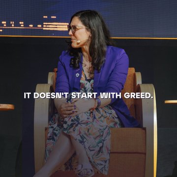 A woman with glasses wearing a blue blazer sits on a stage, with the text overlay reading "IT DOESN'T START WITH GREED." The image accompanies a message about Bitcoin being rooted in personal agency and taking control of one's financial future rather than greed.