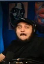A photo of a bearded man wearing a black cap and headphones, appearing to be streaming or recording a video with art on the wall behind him.