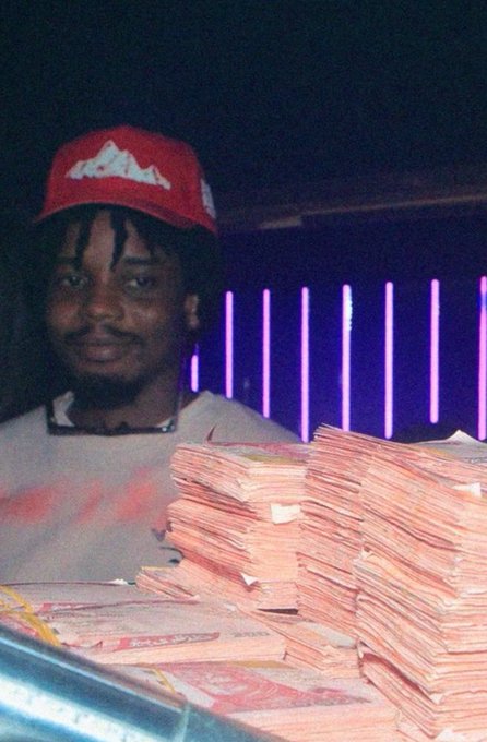A photograph of a smiling young man wearing a red cap, standing behind large stacks of banded cash in a room with purple neon lighting.