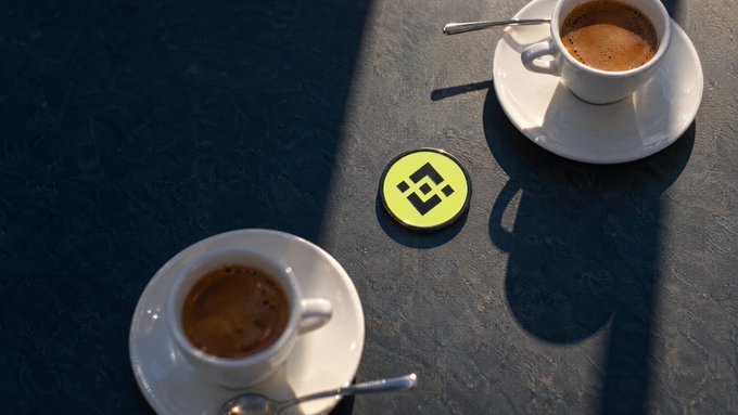Two small espresso cups sit on white saucers on a dark gray surface flanking a round Binance (BNB) coin token in the center.